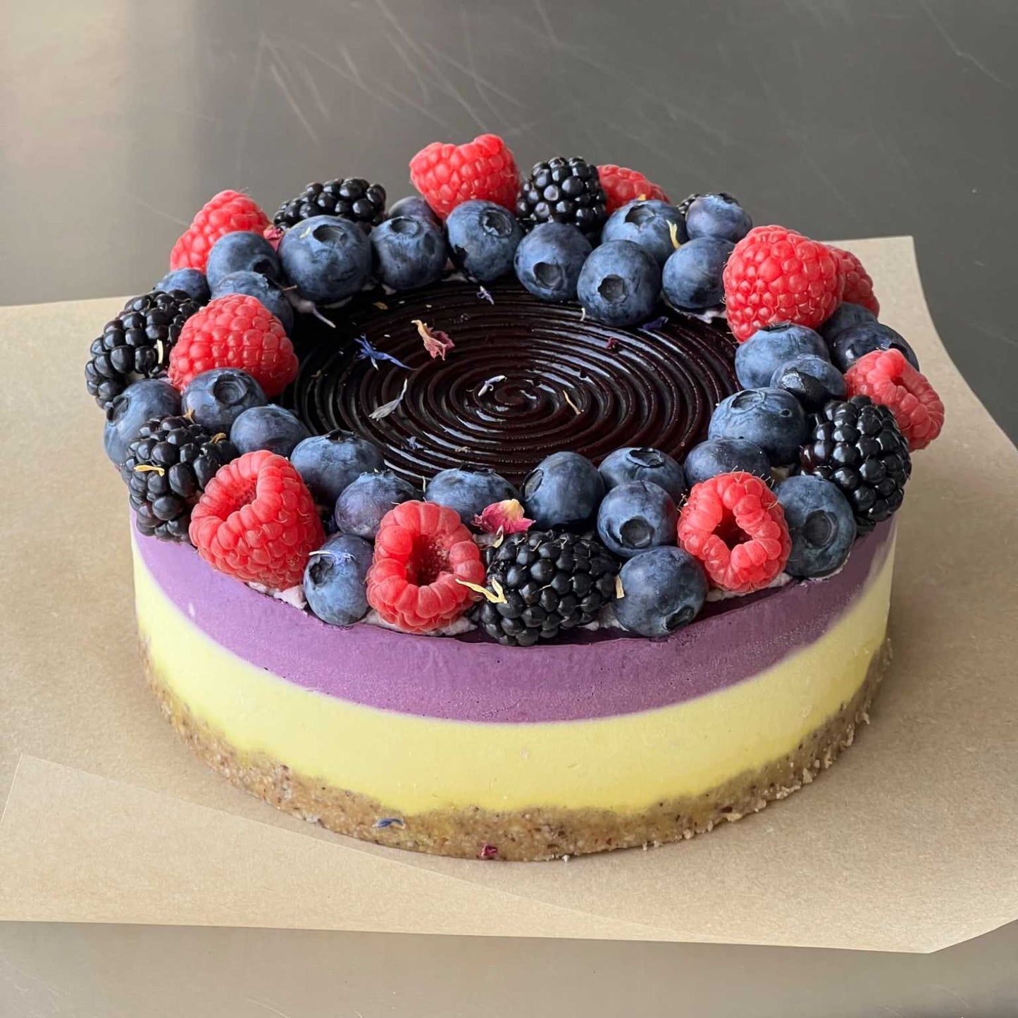 Ein zuckerfreier, glutenfreier und roh-veganer Kuchen mit nussigem Boden, leuchtend gelber und violetter Creme, getoppt mit einer fruchtigen Heidelbeer-Glasur und frischen Himbeeren, Brombeeren und Heidelbeeren – ein gesunder Genuss aus natürlichen Zutaten.