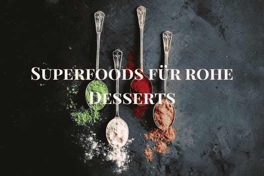 Vier Löffel mit bunten Superfood-Pulvern – grün, rot, weiß und braun – auf dunklem Hintergrund, mit dem Text „Superfoods für rohe Desserts“.