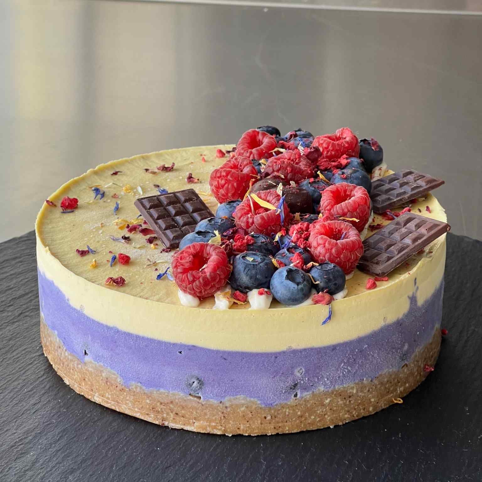 Zuckerfreier Kuchen aus München – ein kunstvoller roh-veganer Genuss mit Heidelbeeren, Mango, nussigem Boden und cremigen Schichten, liebevoll verziert mit frischen Beeren, dunkler Schokolade und Blüten.