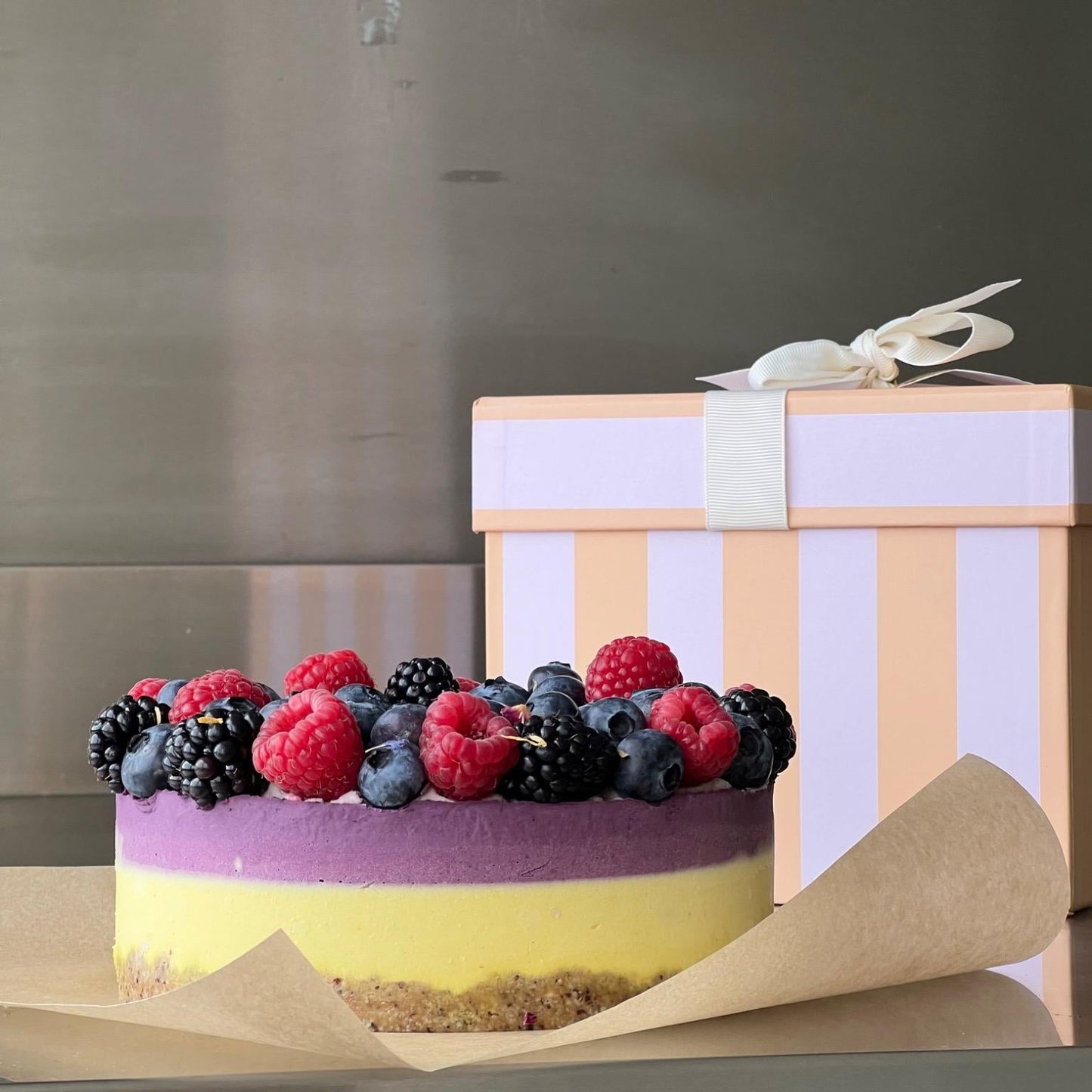 Ein farbenfroher, roh-veganer Kuchen mit nussigem Boden, leuchtend gelber und violetter Creme, belegt mit frischen Himbeeren, Brombeeren und Heidelbeeren. Der zuckerfreie und glutenfreie Kuchen steht auf Backpapier, im Hintergrund eine gestreifte Geschenkbox – ideal für besondere Anlässe.