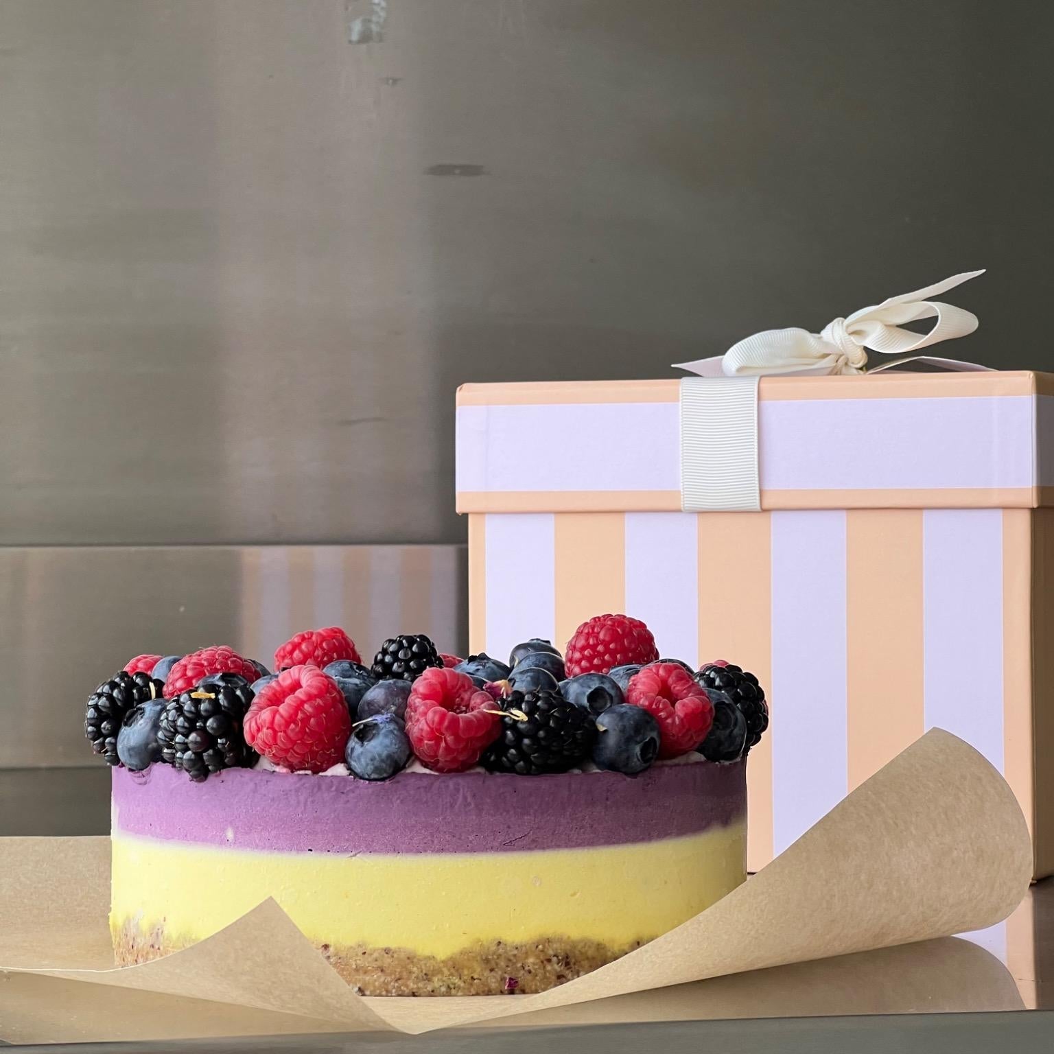 Ein farbenfroher, roh-veganer Kuchen mit nussigem Boden, leuchtend gelber und violetter Creme, belegt mit frischen Himbeeren, Brombeeren und Heidelbeeren. Der zuckerfreie und glutenfreie Kuchen steht auf Backpapier, im Hintergrund eine gestreifte Geschenkbox – ideal für besondere Anlässe.